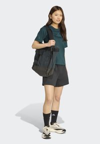 Tmavomodrý tričko, čierne športové šortky, čierne a béžové topánky, nesúci sivý batoh s čiernym dnom a nastaviteľnými popruhmi.