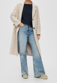 Béžový vlněný kabát s klopovým límcem, zapínáním na knoflíky, rozšířené světle modré džíny a béžové platformové tenisky. Viditelný texturovaný pletený top pod ním.