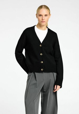 Zwarte gebreide cardigan met een V-hals, voorzien van vijf knoopsluitingen in een contrasterende kleur en geribbelde manchetten, gecombineerd met grijze broeken.