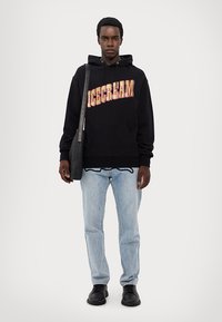 Schwarzer Hoodie mit "ICECREAM" Grafik in Rot und Gelb, kombiniert mit hellblauen Jeans und schwarzen Schuhen, trägt eine schwarze Umhängetasche.