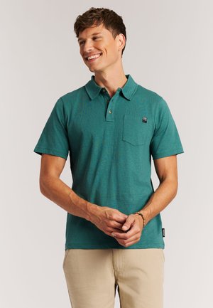 Lachende man die een teal polo met korte mouwen en een borstzak draagt, gecombineerd met beige broek, staand tegen een effen achtergrond.