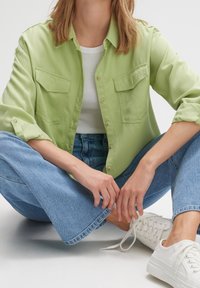 Femme assise jambes croisées portant une chemise verte claire boutonnée, un débardeur blanc côtelé, un jean bleu et des baskets blanches aux lacets défaits.