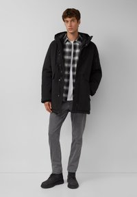 Schwarze Parka-Jacke mit Kapuze, vorderen Knöpfen und Seitentaschen, über einem schwarz-weißen Karohemd, grauen Jeans und schwarzen Stiefeln getragen.