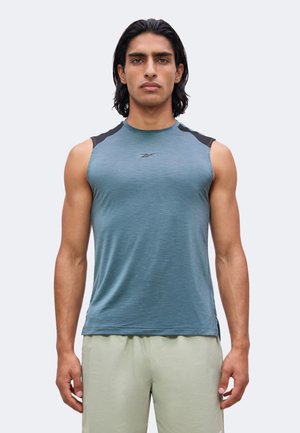 Jeune homme aux cheveux foncés mi-longs porte un maillot de sport bleu sans manches et un short de couleur claire, debout devant un fond uni.