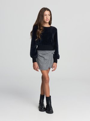 Maglione nero corto e peloso con polsini a coste abbinato a una gonna grigia a righe sottili con fiocco e stivaletti neri alla caviglia.