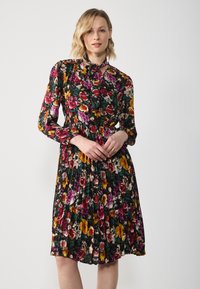 Kukallinen mekko, jossa on musta tausta ja kirkkaat punaiset, keltaiset ja purppurat kukat, pitkät hihat, solmittava kaula-aukko ja laskostettu hame.