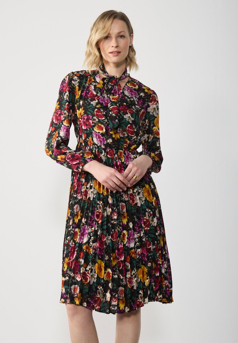 Blumenkleid mit schwarzem Hintergrund, lebendigen roten, gelben und lila Blumen, langen Ärmeln, einem Bindeband am Hals und plissiertem Rock.