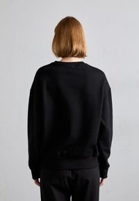 Μαύρη μπλούζα με χαλαρή εφαρμογή, ριμπ μανσέτες και στρίφωμα, καθώς και μια μικρή λεπτομέρεια λογότυπου στο πίσω μέρος του λαιμού, φτιαγμένη από μαλακό βαμβακερό υλικό.