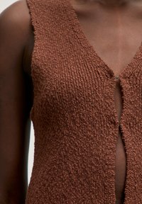 Braun gestricktes Kleidungsstück mit tiefem V-Ausschnitt, strukturierter Oberfläche und Frontverschluss. Der Stoff ist weich und am Ausschnitt leicht offen.