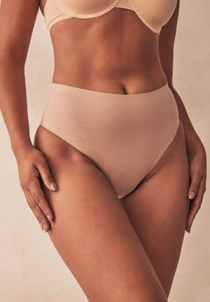 Femeie care poartă lenjerie nude fără cusături, cu talie înaltă și sutien asortat, mâinile odihnindu-se pe coapse, pe un fundal bej simplu.