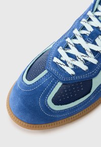 Pantof din piele întoarsă albastră texturată, cu un design complex și accente verde mentă, secțiuni din plasă și șireturi albe pe o talpă gumă.