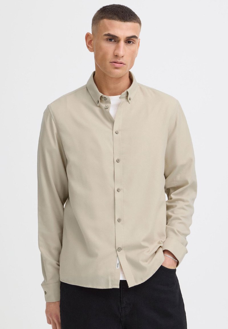 Jeune homme aux cheveux courts portant une chemise beige boutonnée sur un haut blanc, avec une main dans la poche de son pantalon noir.