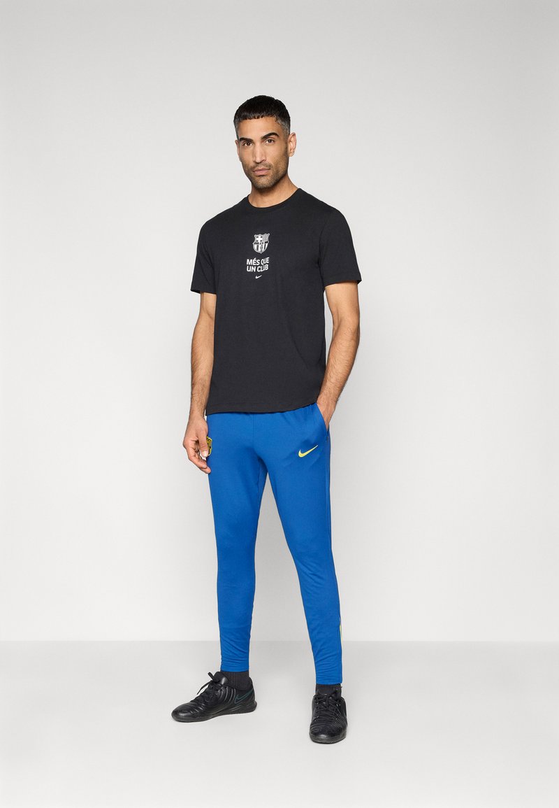 Schwarzes Baumwoll-T-Shirt mit weißem Text und Logo; blaue sportliche Jogginghosen mit gelbem Nike-Swoosh; schwarze Sportschuhe.