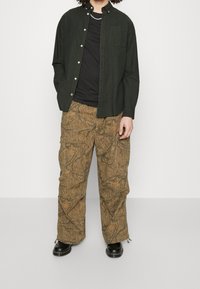 Tummaan vihreä nappipaita, musta t-paita alla ja ruskeat cargo-housut, joissa on teksturoitu lehtikuvio. Mustat kengät viimeistelevät asun.