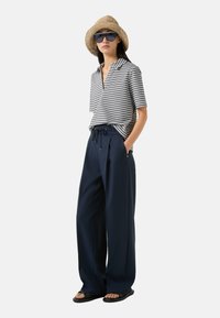 Gestreepte korte mouwen polo in grijs en wit, gecombineerd met marineblauwe wijde broek, een gevlochten strohoed en donkere zonnebril. Casual strandoutfit.