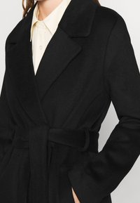 Zwarte wollen wikkelhoed met een riem in de taille, gekartelde kraag en zijzakken. Glad textuur en een getailleerde pasvorm.