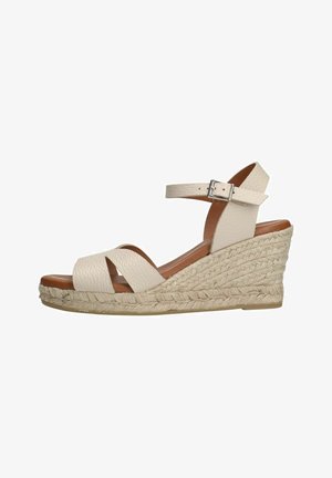 Beige wedge sandal made of textured leather. Features crisscross straps, an ankle strap with a silver buckle, and a jute-wrapped platform.
