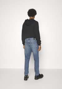 Svart huppartröja och ljusblå jeans med en CK-logga på baksidan. Svarta robusta kängor. Minimalistisk miljö med neutral bakgrund.