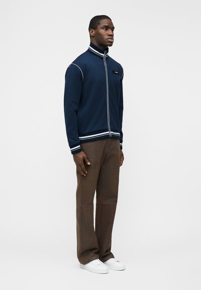 Homme debout de profil portant une veste zippée bleu marine avec des bordures blanches, un pantalon marron et des baskets blanches, sur un fond blanc uni.