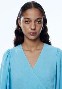 Himmelblaues Kleid mit Wickeldesign, strukturiertem Stoff und gepufften langen Ärmeln. Verfügt über einen tiefen V-Ausschnitt und eine glatte Oberfläche.