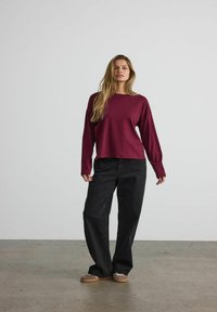 Maglietta a maniche lunghe color bordeaux con vestibilità ampia, abbinata a pantaloni neri a gamba larga. Il tessuto appare morbido; la maglietta presenta un colletto rotondo.