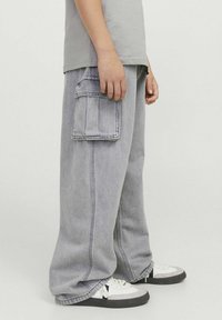 Sivé voľné cargo nohavice so bočnými vreckami, voľný strih a rovné nohavice, v kombinácii s bielymi teniskami. Tkanina vyzerá ako denim a má textúru.