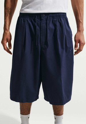 Mężczyzna noszący granatowe szorty do kolan z elastycznym pasem i sznurkiem, białe skarpetki oraz białą koszulę widoczną w talii.