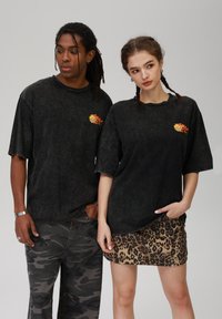 T-shirts de algodão pretos e oversized apresentam um gráfico colorido de sol no lado esquerdo do peito. O modelo masculino usa calças de camuflagem; a modelo feminina usa uma saia com padrão de leopardo.