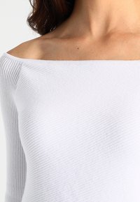 Haut blanc dévoilant les épaules, fabriqué en tissu côtelé. Caractérisé par une surface texturée et une coupe ajustée, mettant en valeur un col sans couture.