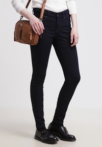 Mørkeblå smale jeans med en subtil glans, sammen med en lys genser og svarte sko. En brun crossover-veske har frontlommer.