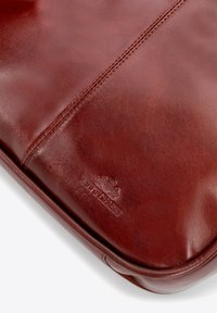 Brązowa skórzana etui na laptopa z gładką teksturą, subtelnymi detalami przeszycia i wytłoczonym logo w dolnym rogu.