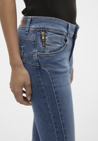 Los jeans de mezclilla azul tienen un corte ajustado, cierre frontal y bolsillos laterales. Costuras distintivas a lo largo de los lados y una etiqueta de cuero en la cintura trasera.