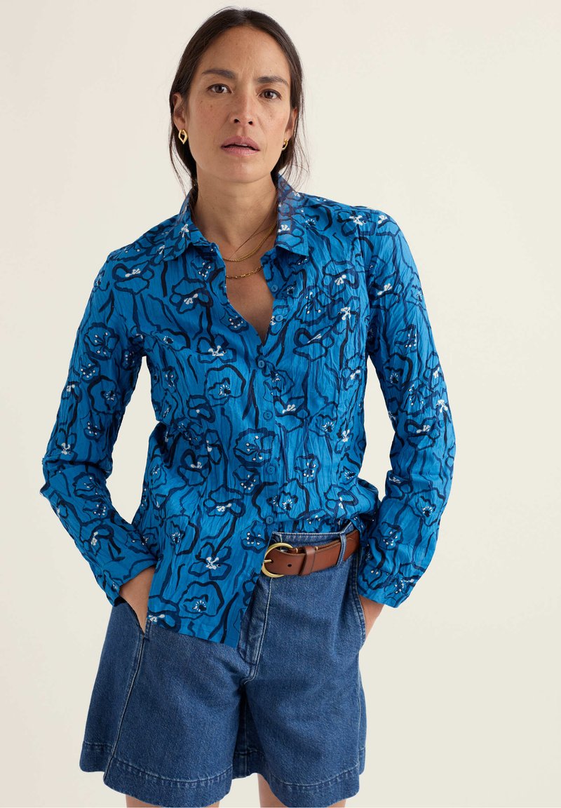 Vrouw draagt een blauw bloemenlangarmshirt in een hoge taille denimshort met een bruine riem, staand met handen in de zakken.