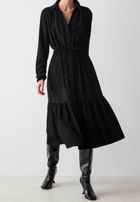 Nainen, joka on pukeutunut pitkähiharaiseen mustaan midi-mekkoon, jossa on vyötäröltä kiristetty malli ja etunapit, yhdistettynä kiiltäviin mustiin polven yli ulottuviin saappaisiin.