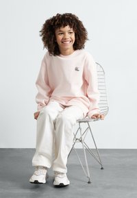 Sudadera rosa claro con cuello redondo y pequeño logo, combinada con pantalones anchos blancos y zapatillas ligeras, sentada en una silla de alambre.