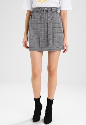 Kobieta ubrana w mini spódnicę w czarno-białą kratę typu pepitka z przewiązanym paskiem, w zestawieniu z czarnymi botkami za kostkę i białym t-shirtem.