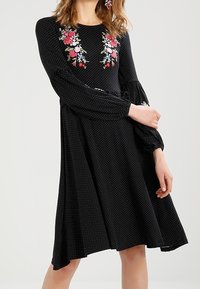 Vestido negro con lunares, de mangas largas y acampanadas, con bordados florales en el pecho en rojo, rosa y azul. Diseño fluido en línea A.