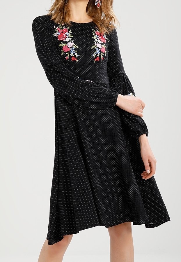 Vestido negro con lunares, de mangas largas y acampanadas, con bordados florales en el pecho en rojo, rosa y azul. Diseño fluido en línea A.