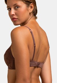 Soutien-gorge en dentelle marron avec bretelles ajustables, motif floral et fermeture à crochet. Le design présente une texture douce et une forme structurée.