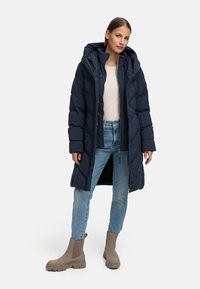 Tumši zils pūkains mētelis ar kapuci, kas aprīkots ar rāvējslēdzēju un pogu aizdari, ar quiltējošu dizainu un priekšējām kabatām; apvienots ar gaiši ziliem džinsiem un bēšiem potītes zābakiem.