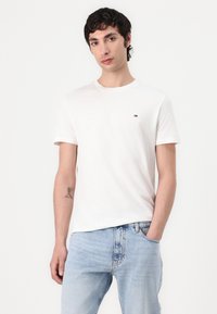 T-shirt en coton blanc avec un col rond et des manches courtes, présentant un petit logo multicolore sur la poitrine. Assorti à un jean bleu clair.