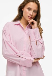 Šviesiai rožinė mygtukais užsegama marškinėliai su ilgomis rankovėmis, minkšta medžiaga, su vieno mygtuko rankogaliais ir klasikinis apykaklė. Lengvas blizgesys ir laisvas siluetas.