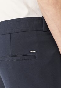 Marineblauwe broek met een gladde textuur, voorzien van een achterzak en metallic logo-accent. Naaddetailing langs de tailleband.