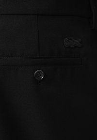 Black tailored trousers made of textured fabric featuring a side pocket, a black button, and a small black crocodile logo.