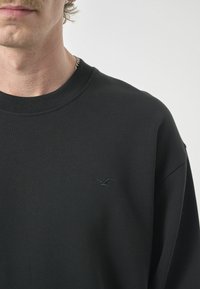 Schwarzer Sweatshirt mit Rundhalskragen, ausgestattet mit einem strukturierten Stoff und einem kleinen gestickten Logo auf der linken Brust.