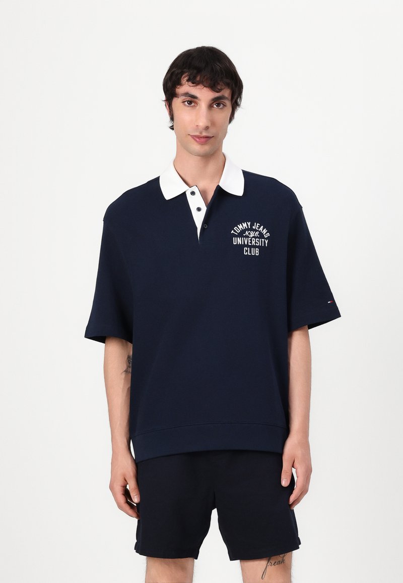 Sötétkék polo ing fehér gallérral és rövid ujakkal, bal mellen hímzett "Tommy Jeans University Club" logóval.