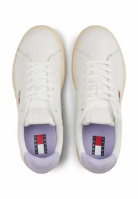 Witte leren sneakers met platte veters, beige zolen en een paarse binnenkant. Een klein logo in rood, wit en blauw is zichtbaar.