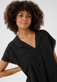 Black shirt with short sleeves, featuring a v-neck, two front pockets, and lace trim on the sleeves. Smooth texture and loose fit.