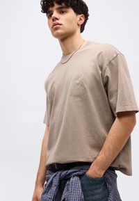 Νέος άνδρας με σγουρά μαλλιά, που φοράει μπεζ γραφικό t-shirt, ασημένια αλυσίδα, μπλε τζιν και μια δεμένη καρό πουκάμισο στη μέση, με φωτεινό φόντο.