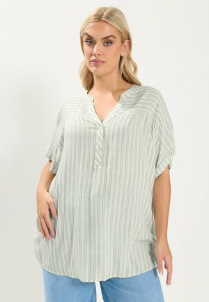 Donna bionda che indossa una camicetta a maniche corte con righe verticali verde chiaro e bianco e jeans blu, in piedi con una mano in tasca contro uno sfondo semplice.